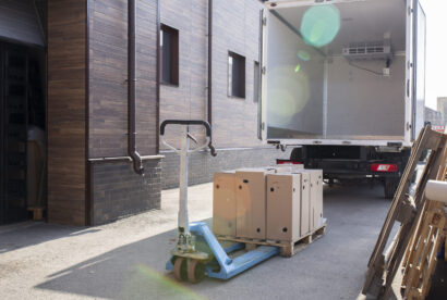 A large box truck backed into a loading bay being loaded with wooden pallets by a forklift.