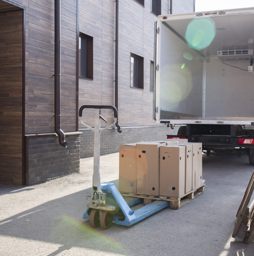 A large box truck backed into a loading bay being loaded with wooden pallets by a forklift.